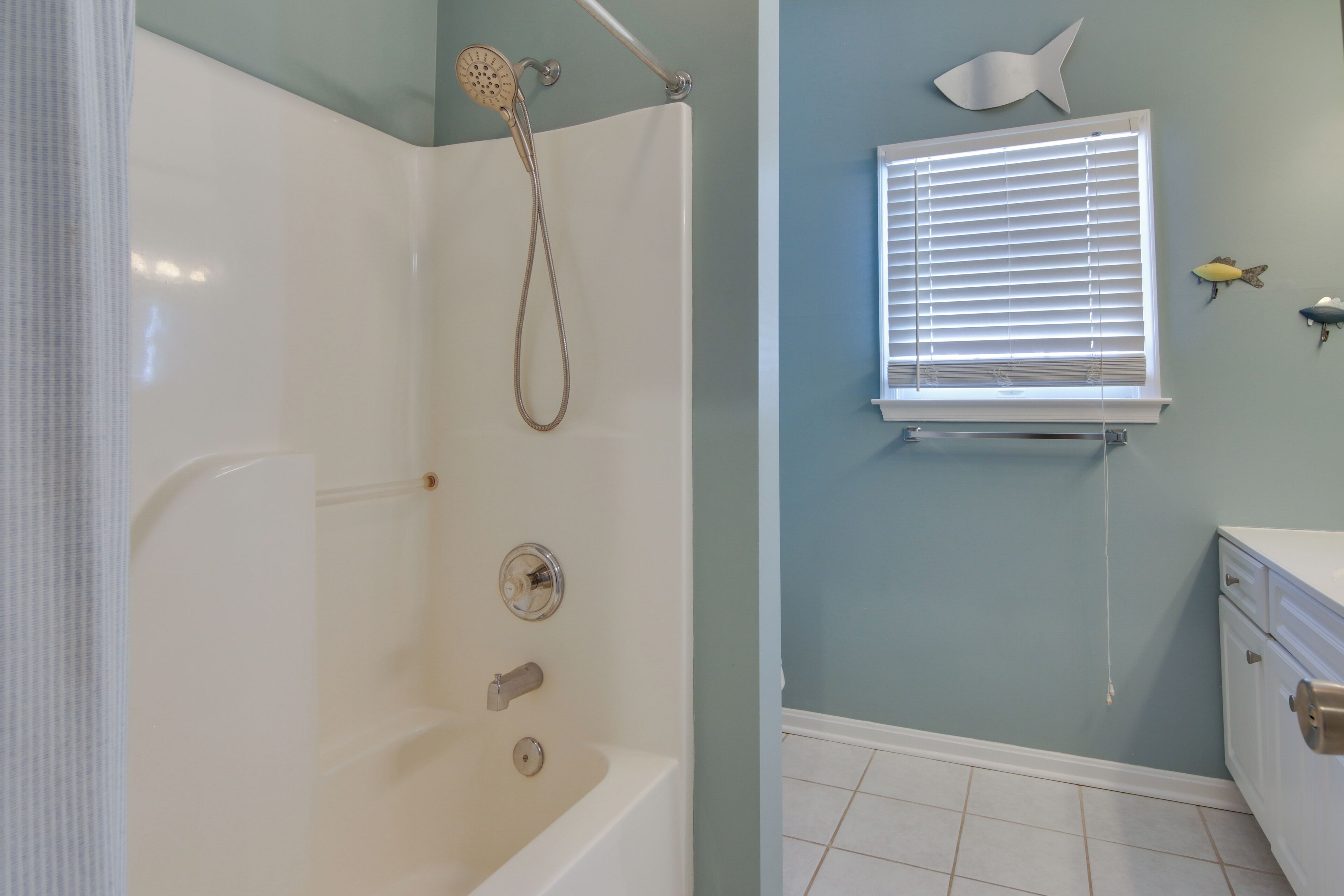 A bathroom with a bathtub, a shower, a window with blinds, and a fish-themed wall decoration.