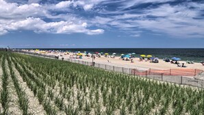 A beach with rows of tall grass in the foreground, a sandy beach with umbrellas and people, and the ocean in the background. - Bay-view Retreat w/ 5 Balconies in Bethany Beach! (Bethany Beach)