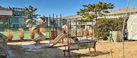 Un parque infantil con toboganes y estructuras para escalar, rodeado por una zona vallada y edificios al fondo.