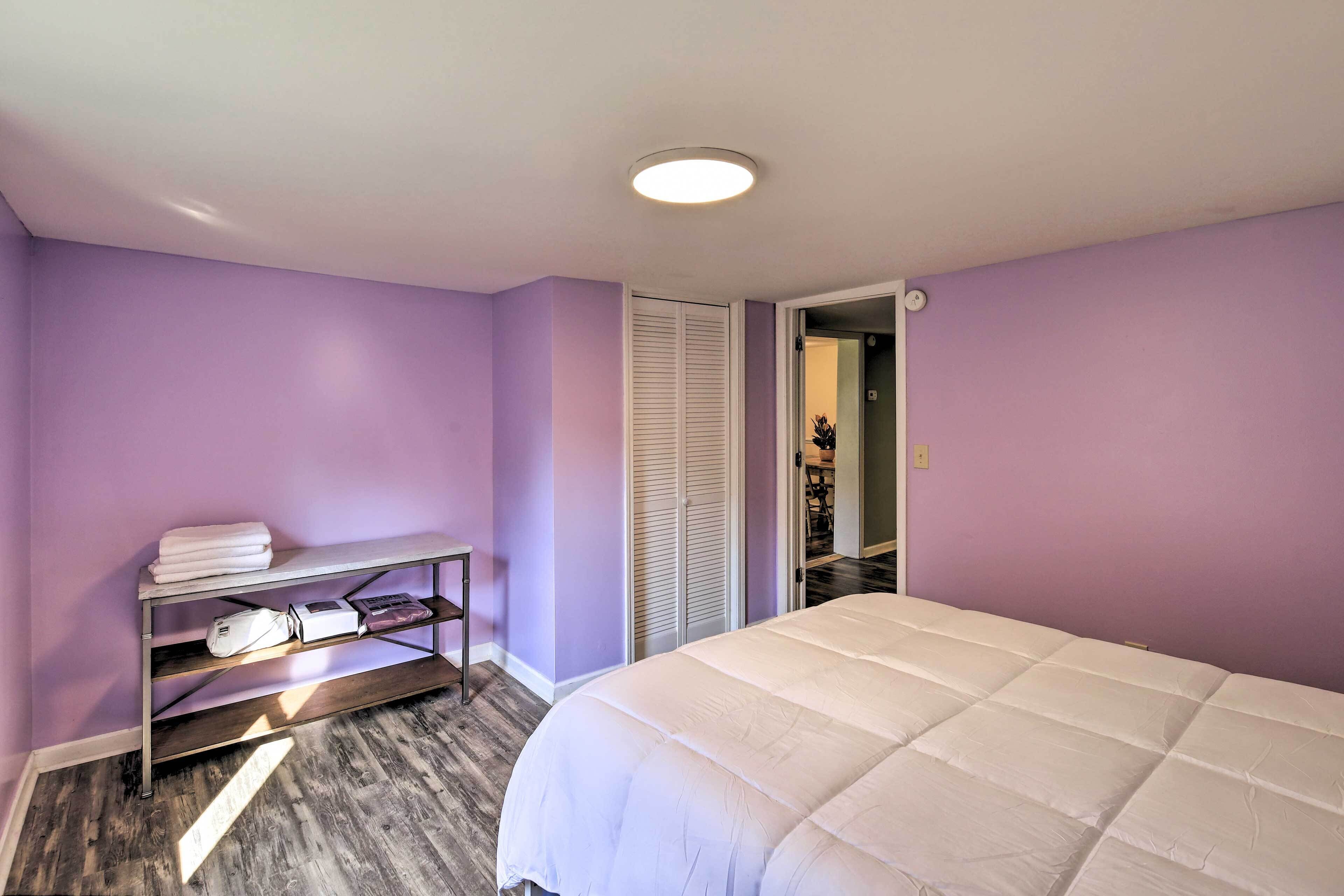 A bedroom with purple walls, a white bed, a wooden floor, and a metal shelf with towels and a bag.