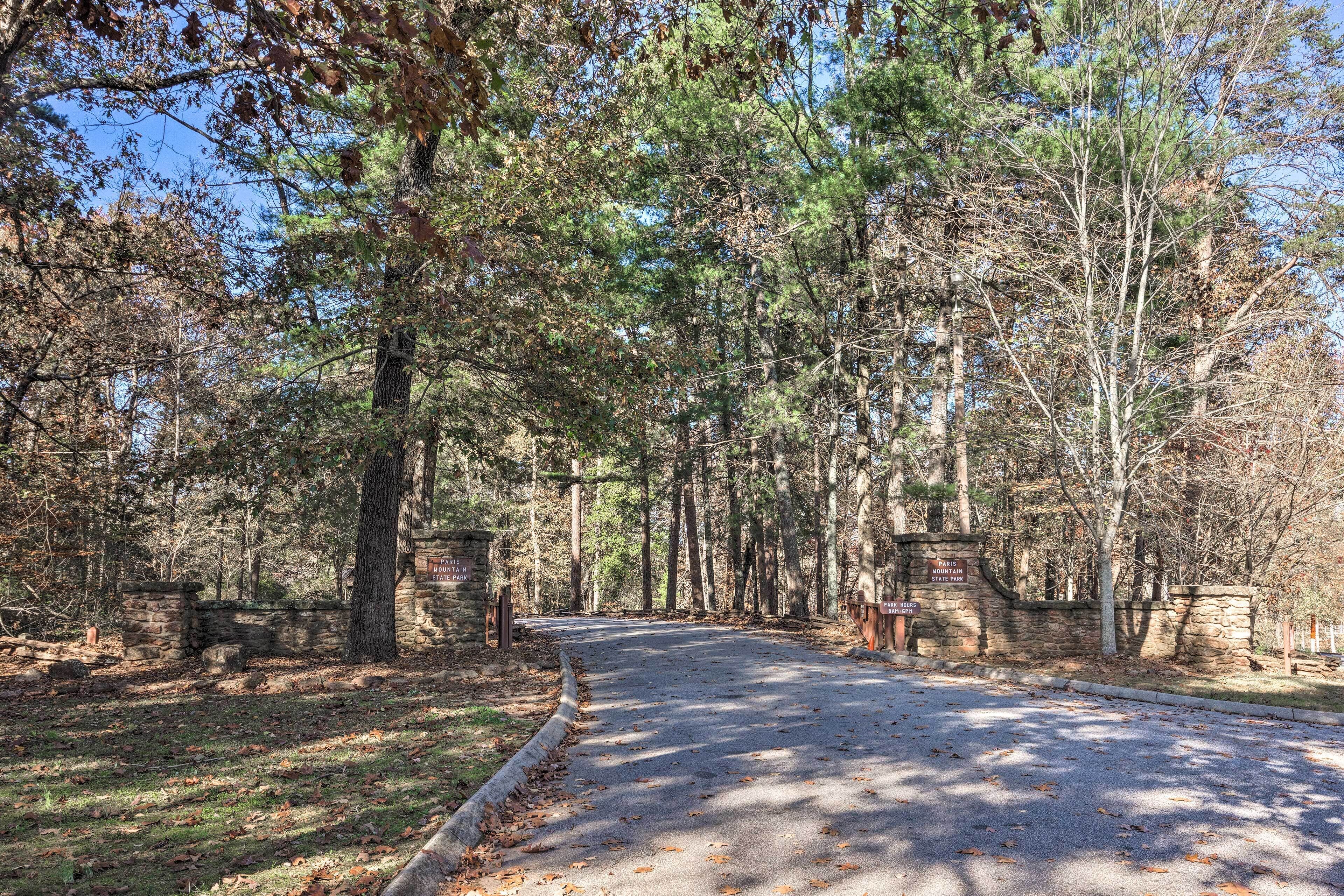 A paved road leading into a wooded area with stone walls and signs indicating park hours.