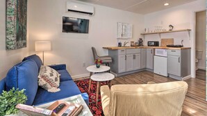 A compact living space with a sofa, a small round table, a wicker chair, a kitchen area with a microwave and cabinets, and a TV mounted on the wall. - Claires Cottage ~ 4 Mi to Downtown Greenville! (Greenville)