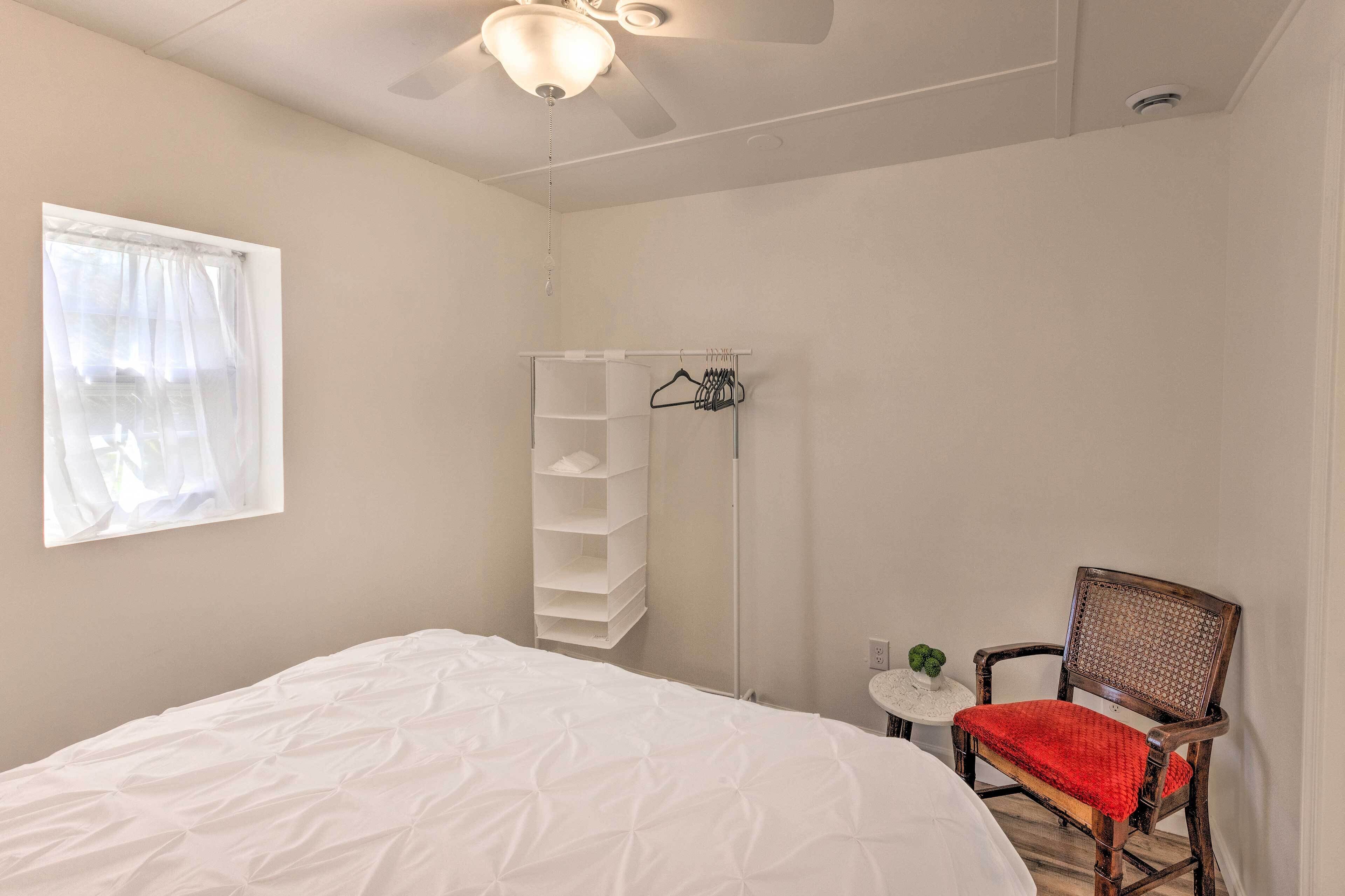 A bedroom with a bed, a chair, a small table, a shelf unit, and a window with sheer curtains.