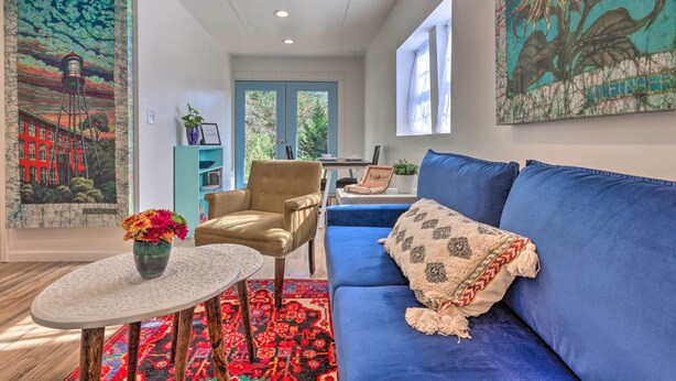 A living room with a blue sofa, a round coffee table with a vase of flowers, a patterned rug, and a chair. There are paintings on the walls and a window with curtains. - Claires Cottage ~ 4 Mi to Downtown Greenville! (Greenville)