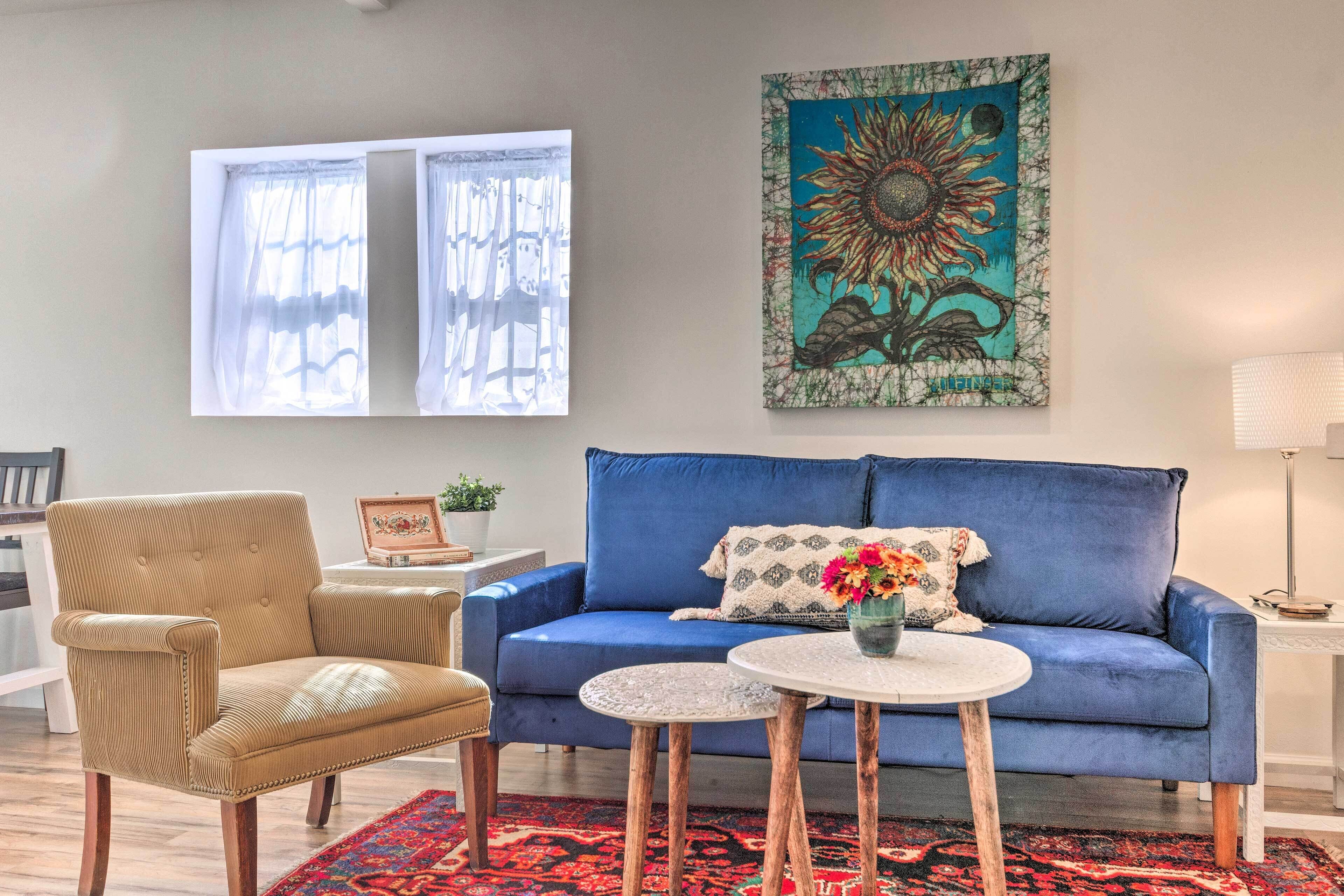 Una sala de estar con un sofá azul, un tapete estampado, una silla de madera, una mesita y un cuadro grande en la pared.