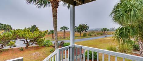 A covered porch with a metal chair, white railing, and a view of a grassy landscape with palm trees and a road.