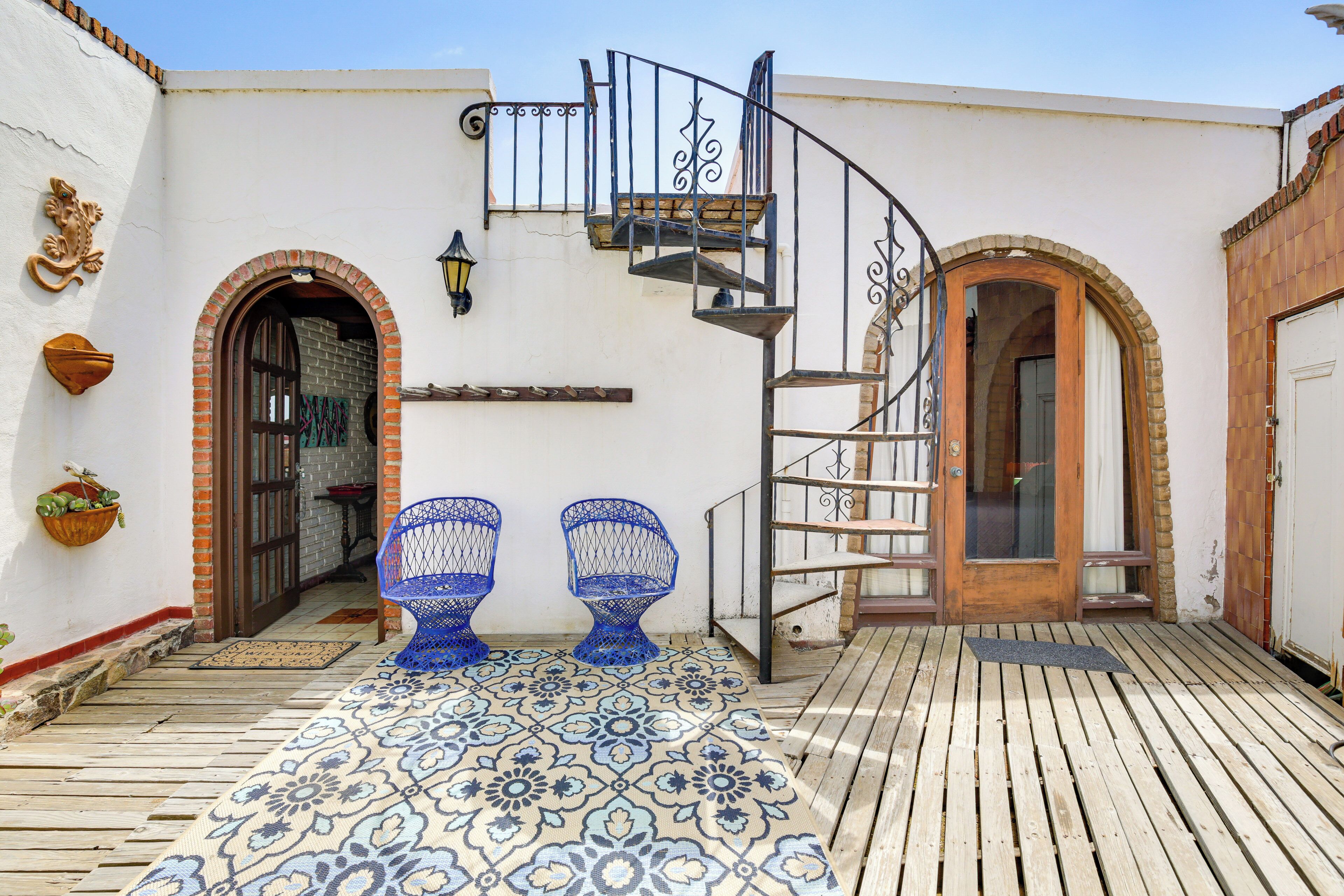 A courtyard with a wooden deck, two blue chairs, a spiral staircase, and a white building with arches and a door.