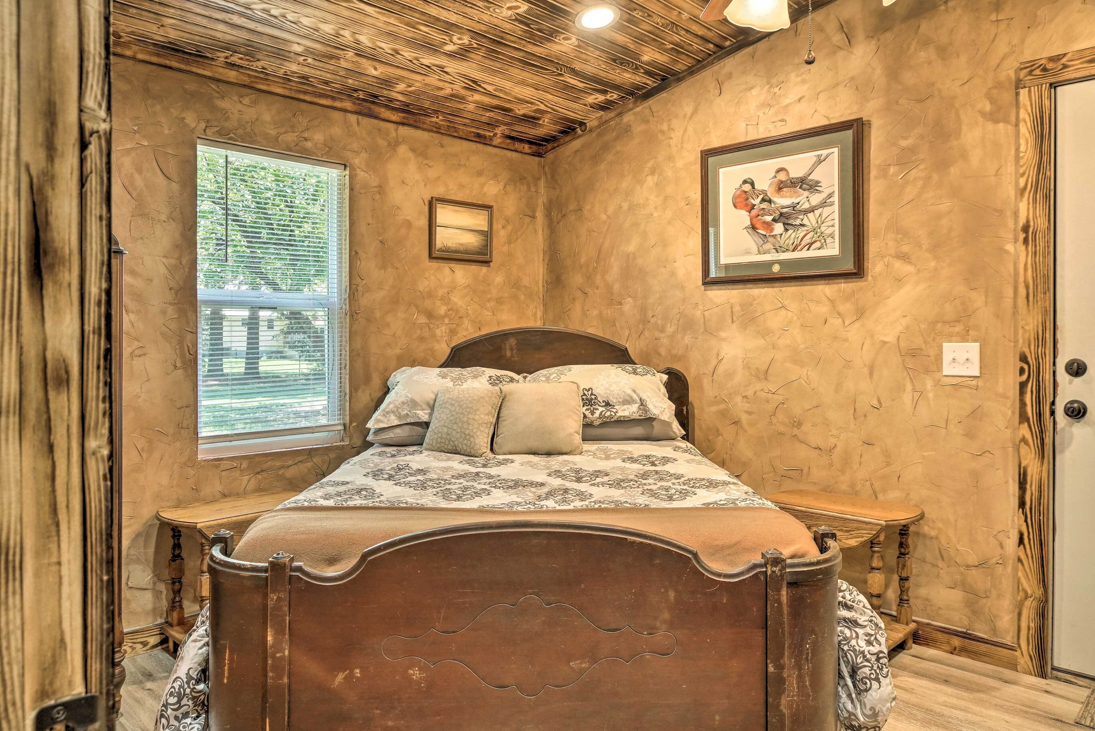 Una camera da letto con soffitto in legno, un letto con lenzuola fantasia, una finestra con persiane e un quadro appeso al muro.