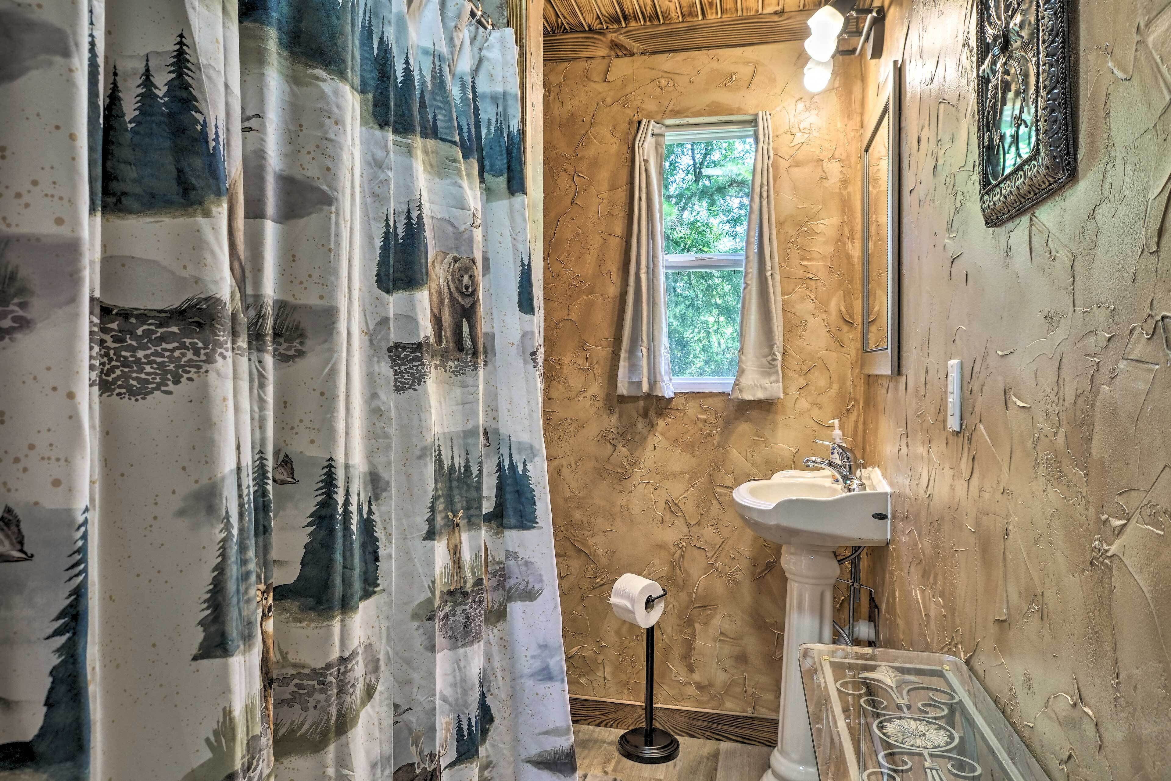A bathroom with a rustic design, featuring a wooden wall, a window, a sink, and a shower curtain with forest-themed prints.