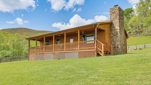 A wooden cabin with a stone chimney, a covered porch, and a view of a green hillside. - Fish, Hike, Hunt: Secluded Cabin in Doe Hill! (Doe Hill)