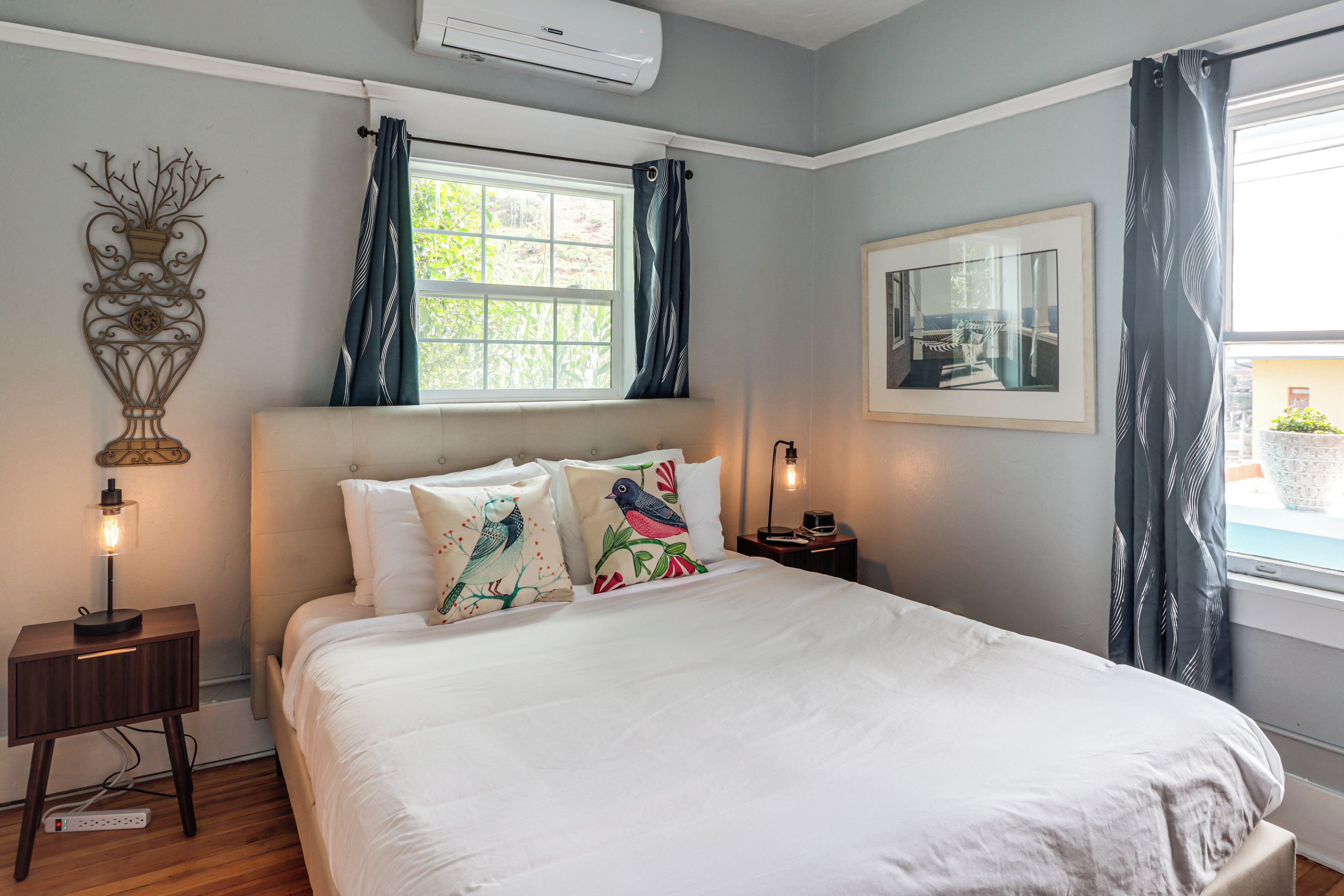 A bedroom with a bed, a nightstand, a lamp, a wall-mounted air conditioner, a window with curtains, and a framed picture on the wall.