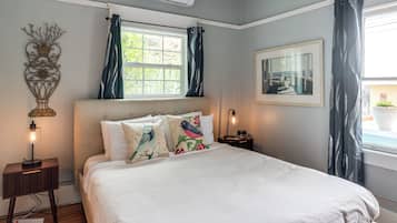 A bedroom with a bed, a nightstand, a lamp, a wall-mounted air conditioner, a window with curtains, and a framed picture on the wall.