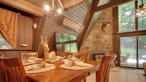 Uma sala de jantar com uma mesa de madeira posta para quatro pessoas, cercada por cadeiras de madeira. Uma lareira de pedra é visível ao fundo.
