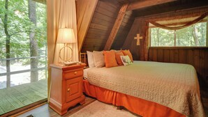 Um quarto com teto de madeira, uma cama com colcha, uma mesa de cabeceira, um abajur e uma janela com uma cruz na parede.