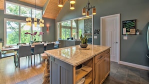 A modern kitchen with granite countertops, wooden cabinets, and a large island. There are multiple pendant lights, a large window, and a view of the outdoors. - Grand Chalet w/ Hot Tub - Near Wintergreen Resort (Nellysford)