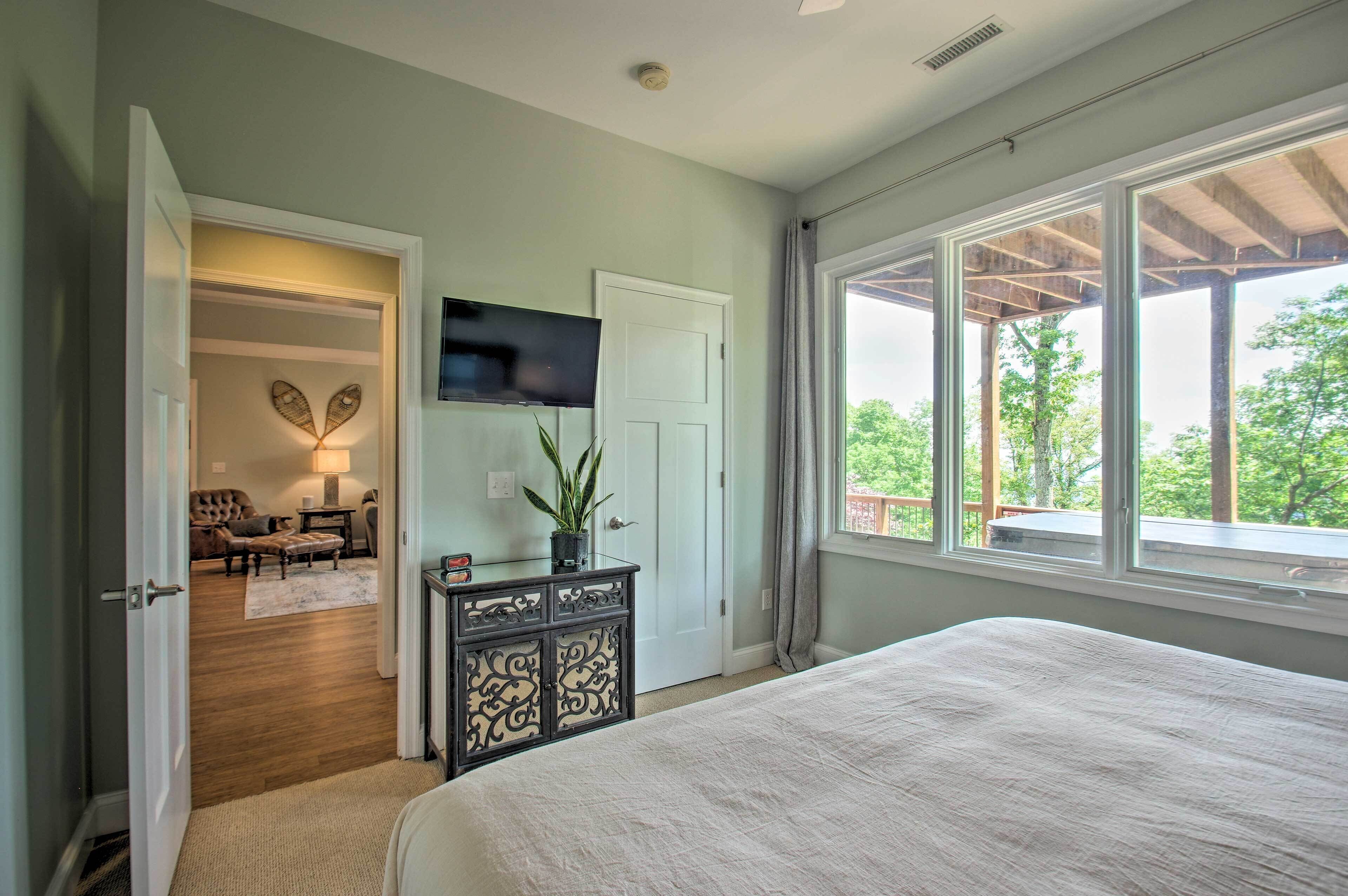 A bedroom with a large bed, a TV mounted on the wall, and a view of the outdoors through a large window.