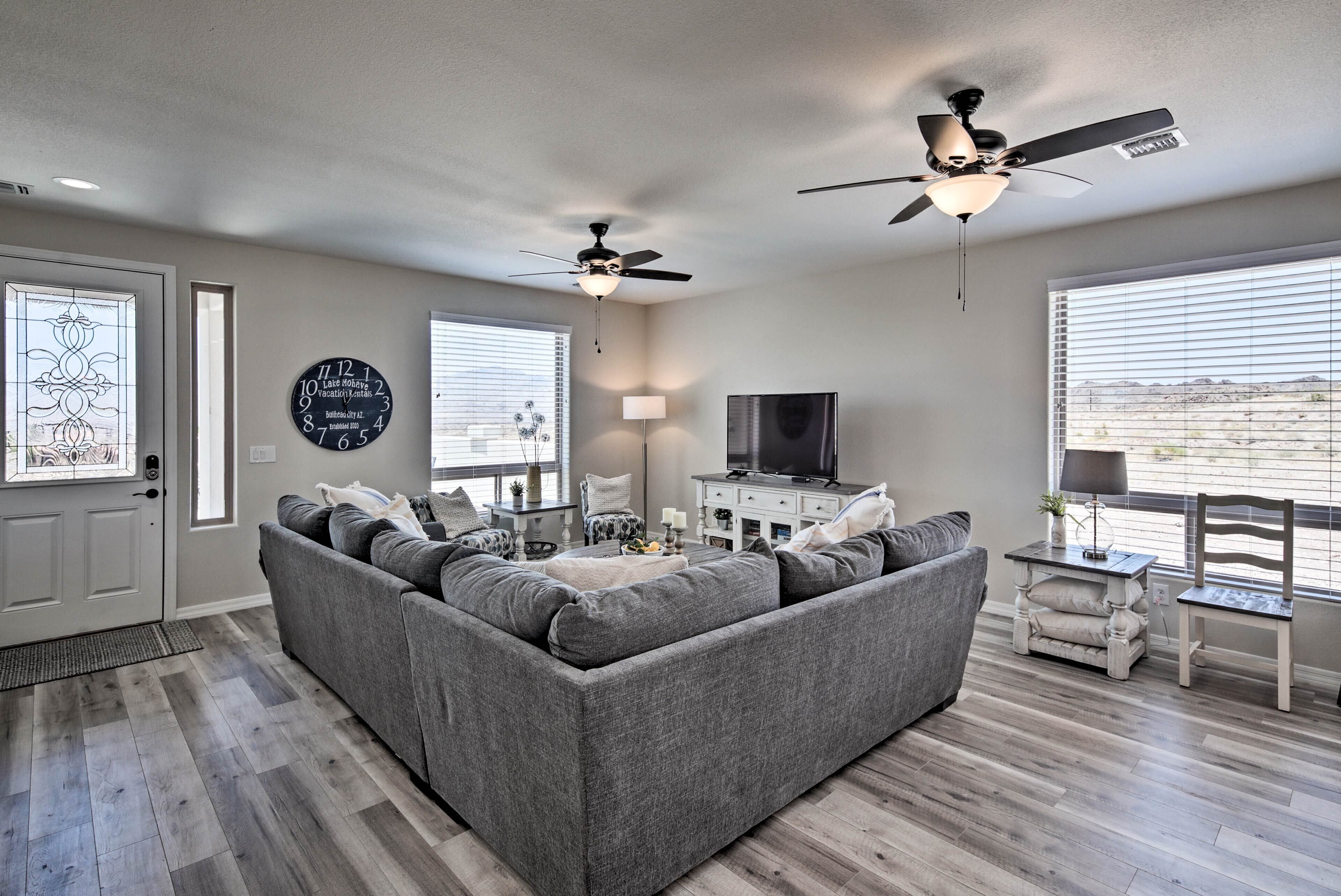 A living room with a grey sectional sofa, two ceiling fans, a TV, and a window with blinds.