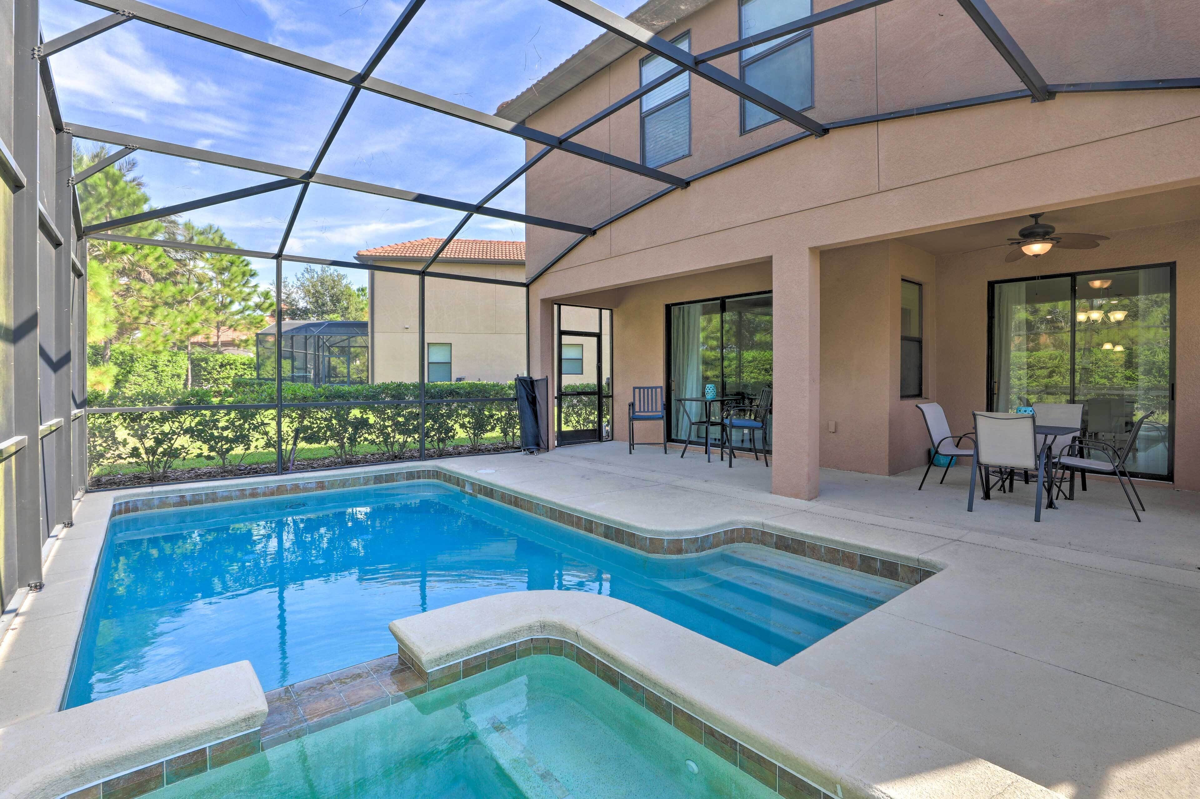 A covered outdoor swimming pool with a glass enclosure, adjacent patio area with chairs and a table, and a view of the surrounding greenery.