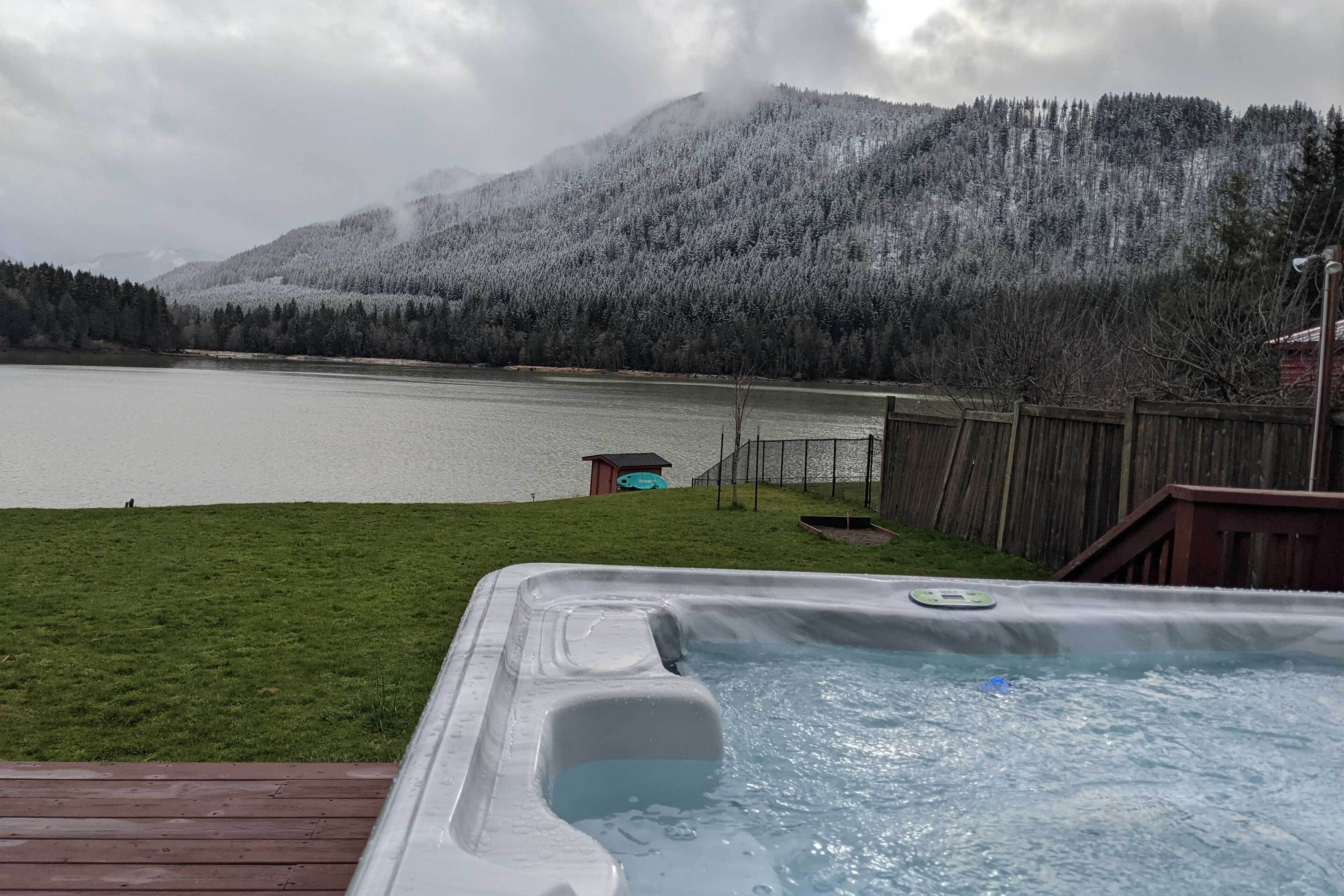 A hot tub on a wooden deck with a view of a lake and snow-covered mountains.