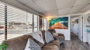 Un salón moderno con un sofá seccional, un ventanal con persianas y un mural de un muelle.