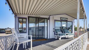 Un área de estar al aire libre techada con dos sillas blancas y una mesa, un cartel que dice 'BEACH' y un letrero de 'BIENVENIDOS' en la puerta.