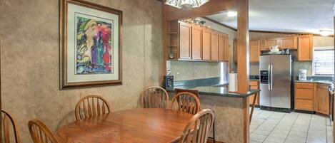 A dining area with a wooden table and chairs, a framed artwork on the wall, and a kitchen with wooden cabinets and a stainless steel refrigerator.