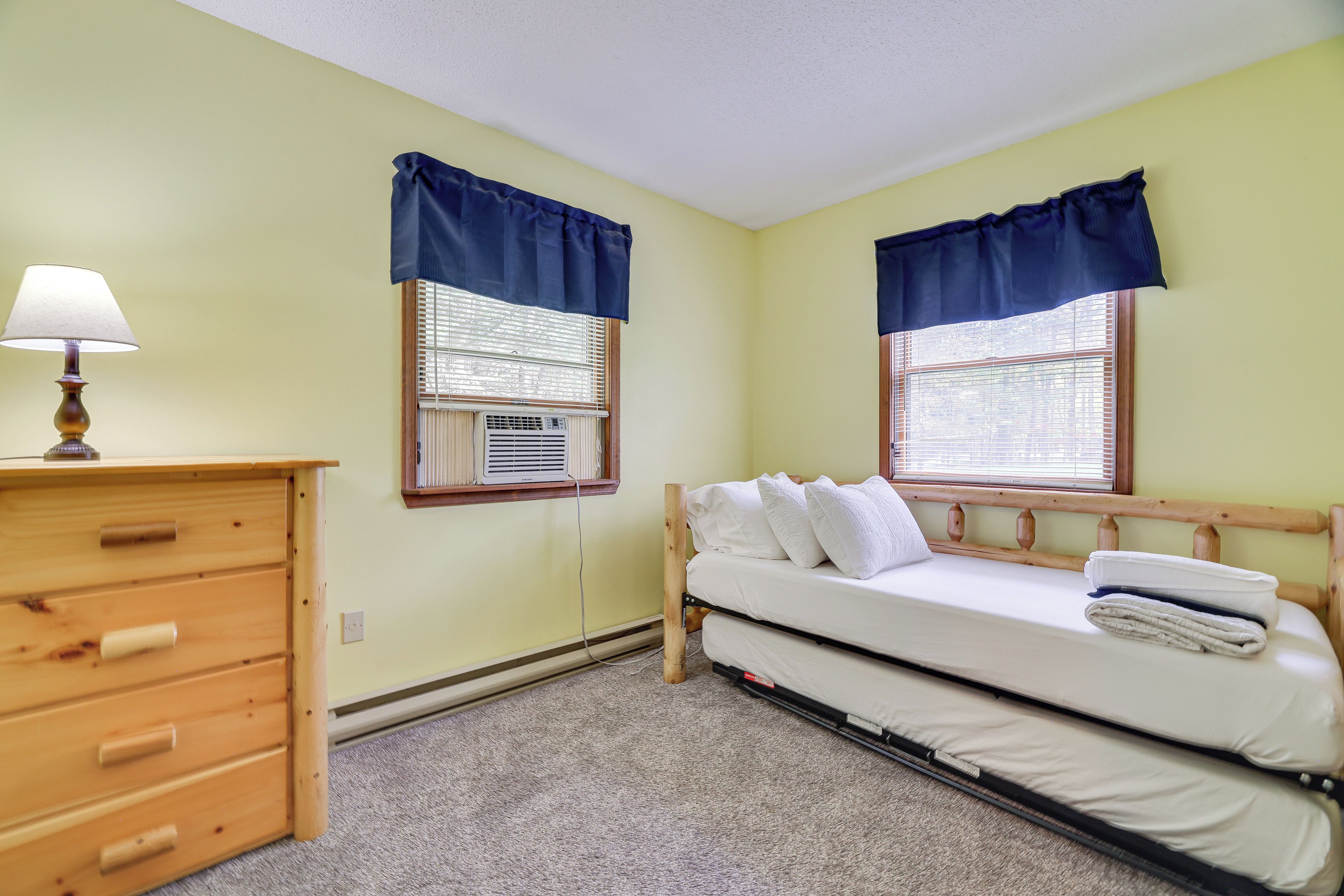 A small, single-bed room with a wooden dresser, two windows with blue valances, and an air conditioning unit.