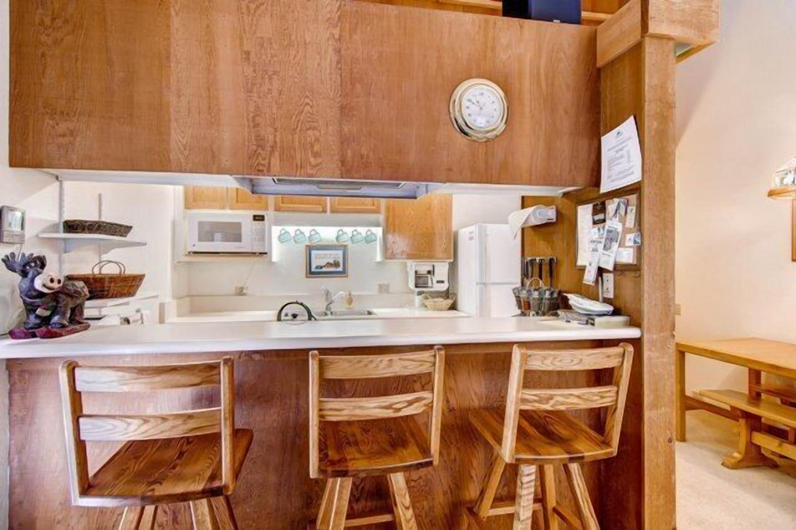 Een keuken met houten kastjes, een wit aanrecht, een magnetron en een klok. Er staan drie houten krukken en een houten bank.