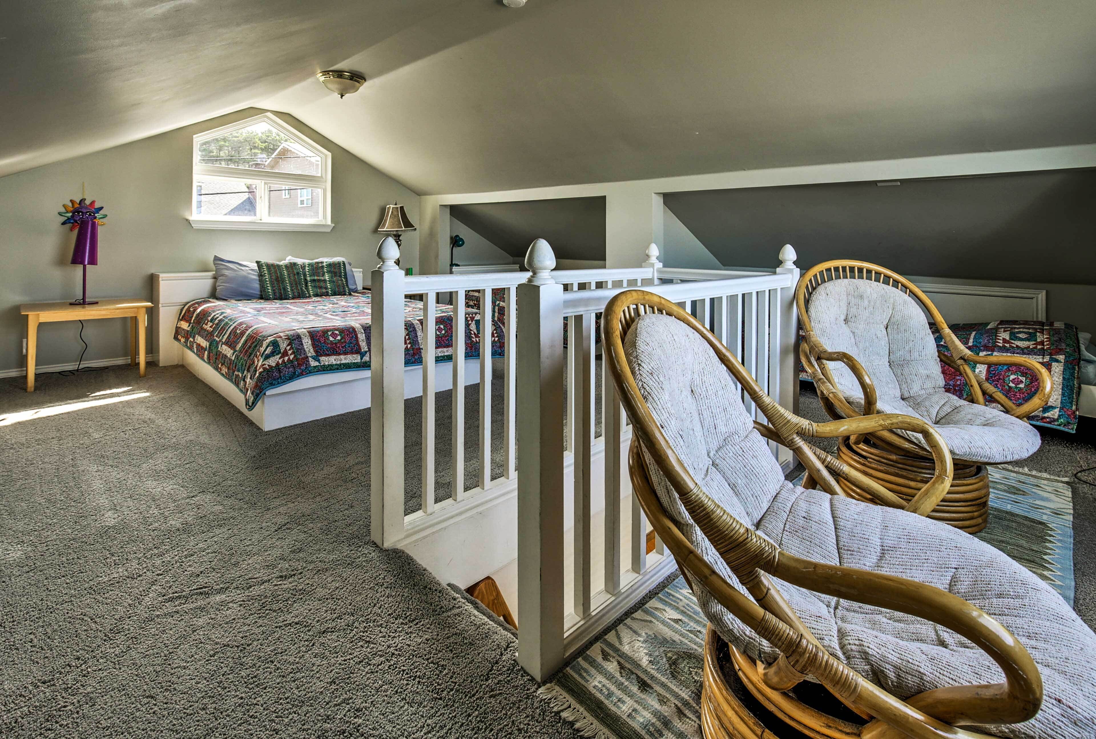 Ein Schlafzimmer mit schrägen Wänden, einem Bett mit farbenfroher Steppdecke, zwei Korbstühlen, einem kleinen Tisch mit Lampe und einem Fenster mit Blick auf ein Haus.