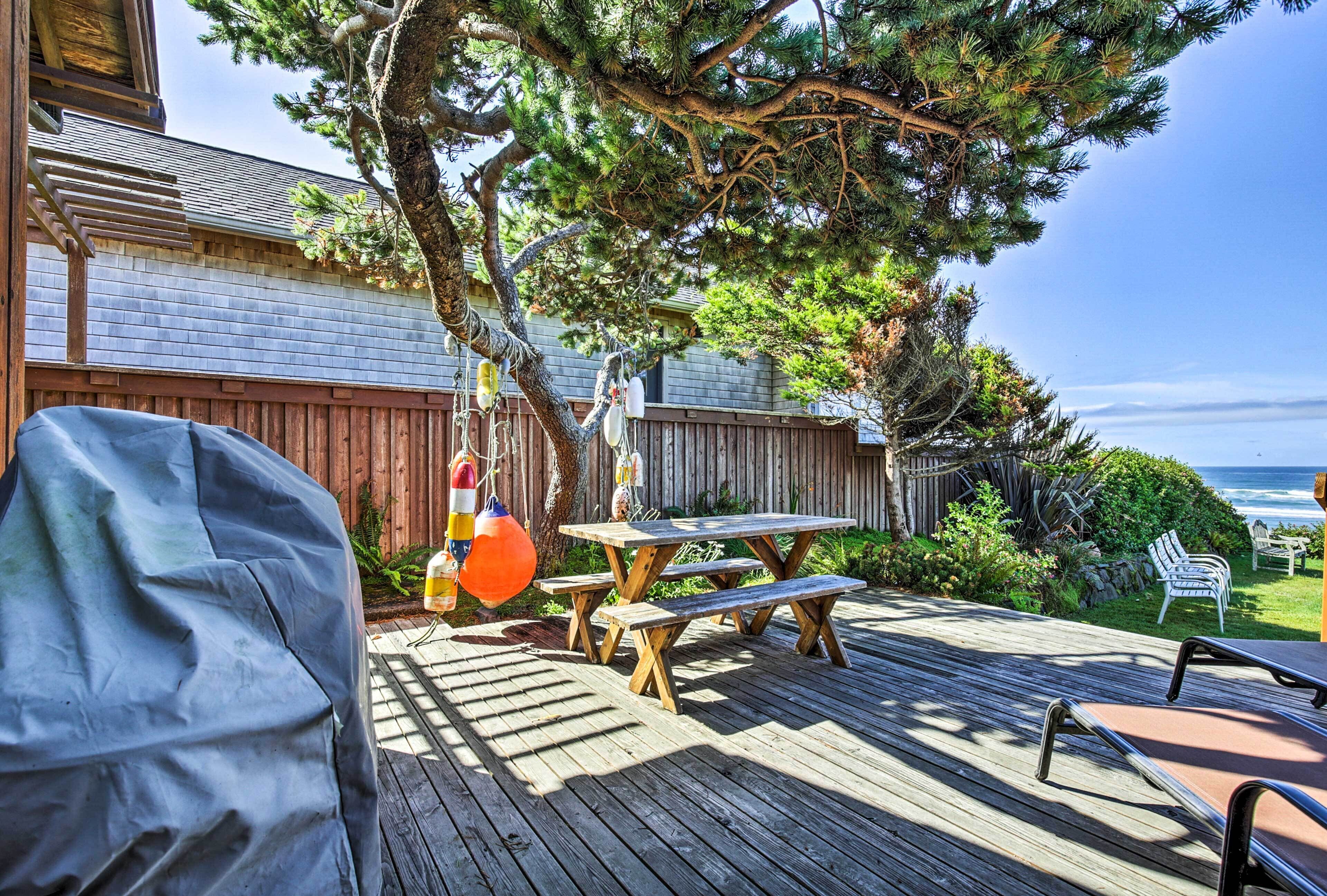 Une terrasse en bois avec des tables de pique-nique et un barbecue, donnant sur une cour arrière bordée d’une clôture en bois et offrant une vue sur l’océan.