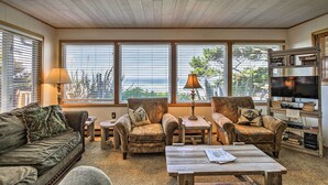 Un salon chaleureux avec un canapé, deux fauteuils, une table basse et un meuble pour la télévision, sur lequel sont posés des livres. De grandes fenêtres offrent une vue sur l’extérieur.