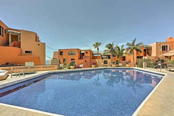 A swimming pool with a tiled edge, surrounded by buildings with a uniform architectural style, palm trees, and a clear sky.