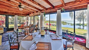 A dining area with a table set for four, surrounded by wicker chairs, a ceiling fan, and large windows overlooking a body of water.
