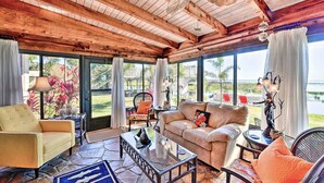 A spacious living room with a stone floor, wooden ceiling beams, and large windows offering a view of the outdoors.