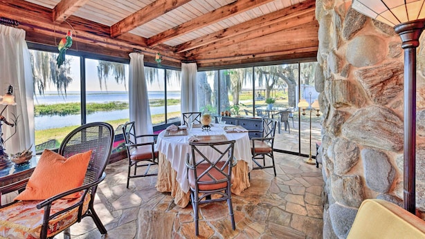A covered outdoor dining area with stone flooring, wooden beams, and large windows overlooking a body of water.