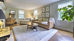 A living room with a sofa, coffee table, and a large window with blinds. - Historic Essex Home w/ Large Yard Near Downtown! (Essex)