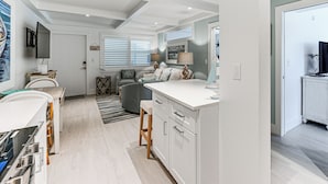 A modern kitchen with white cabinetry, a dining area with a table and chairs, and a living area with a sofa and a TV. - Coastal Downtown Naples Condo - Walk to the Beach! (Naples)