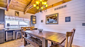 A wooden dining table with benches, a chandelier, and a kitchen area with a refrigerator and sink.
