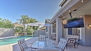 An outdoor patio with a glass table and chairs, a pool, and a TV mounted on the wall. - Arizona Retreat w/ Heated Pool, Fire Pit & Grill! (Surprise)