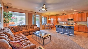 A spacious living room with a large sectional sofa, a glass coffee table, and a dining area with a table and chairs. The kitchen features wooden cabinets and a bar area with stools. - Arizona Retreat w/ Heated Pool, Fire Pit & Grill! (Surprise)