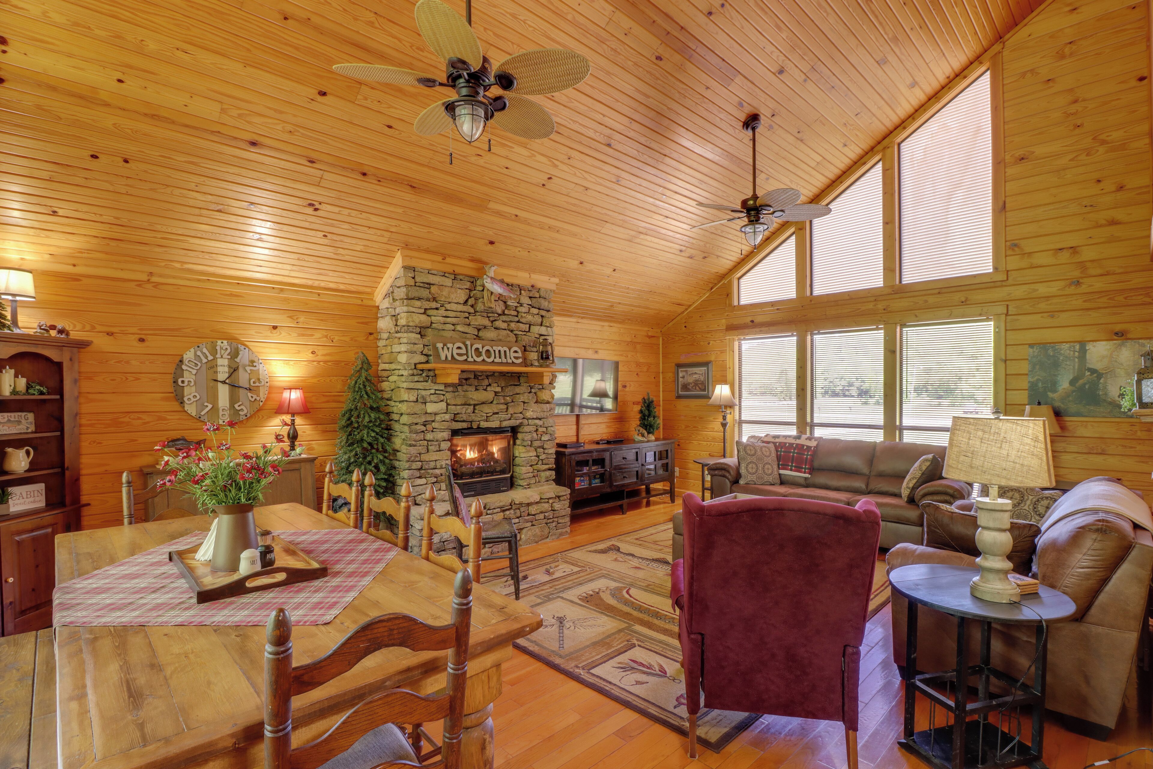 A cozy cabin interior with a stone fireplace, wooden ceiling, and large windows.