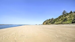 A wide sandy beach with footprints, a clear blue sky, and a forested hillside in the background.
