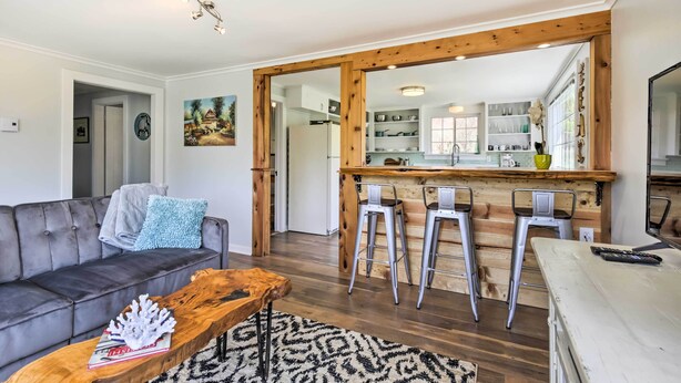 A modern living room with a grey sofa, a wooden coffee table, and a kitchen area with a wooden bar and bar stools.
