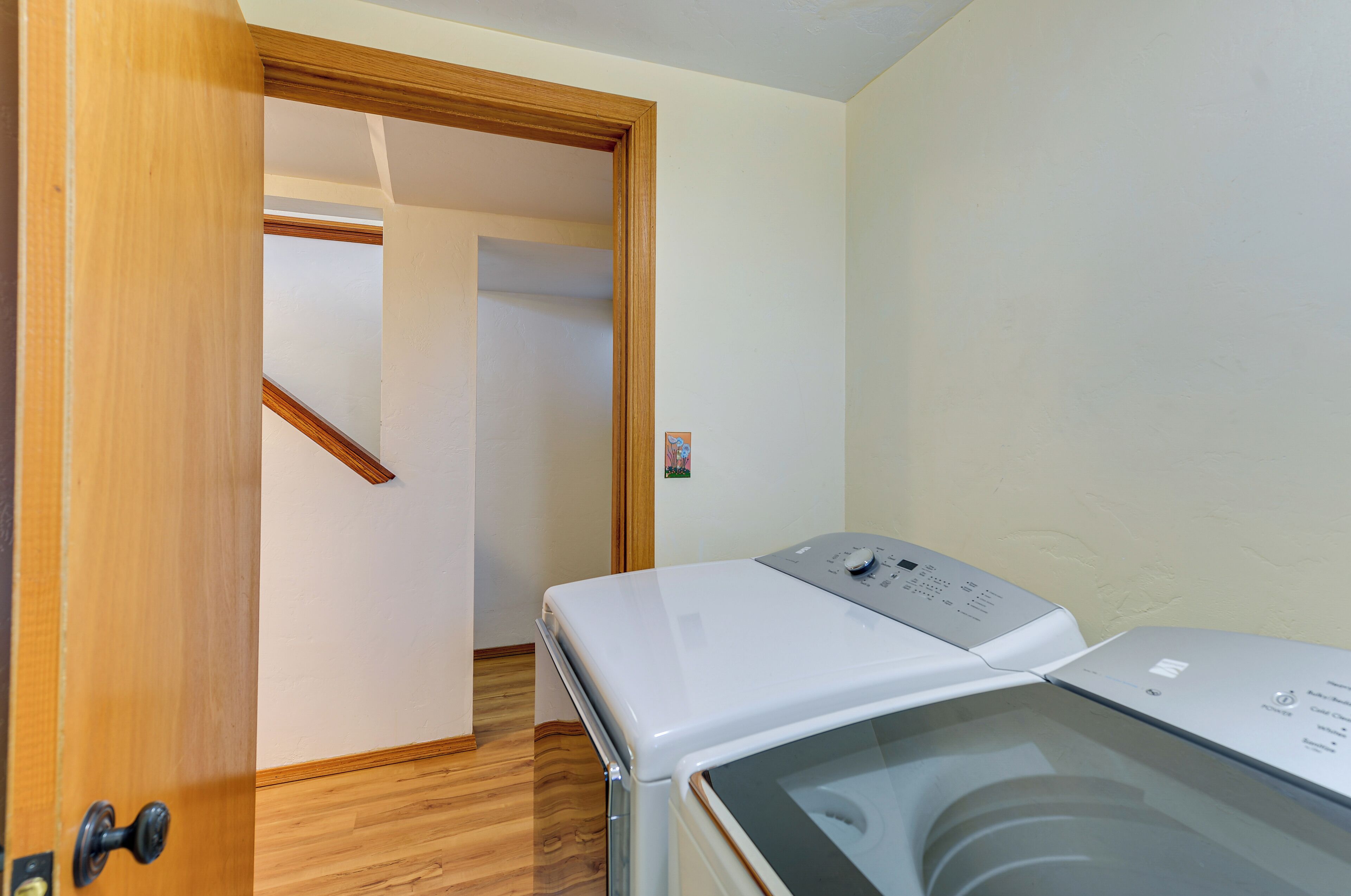 Une buanderie équipée d’un lave-linge et d’un sèche-linge à chargement frontal, avec un plancher en bois et une porte donnant sur une autre pièce.
