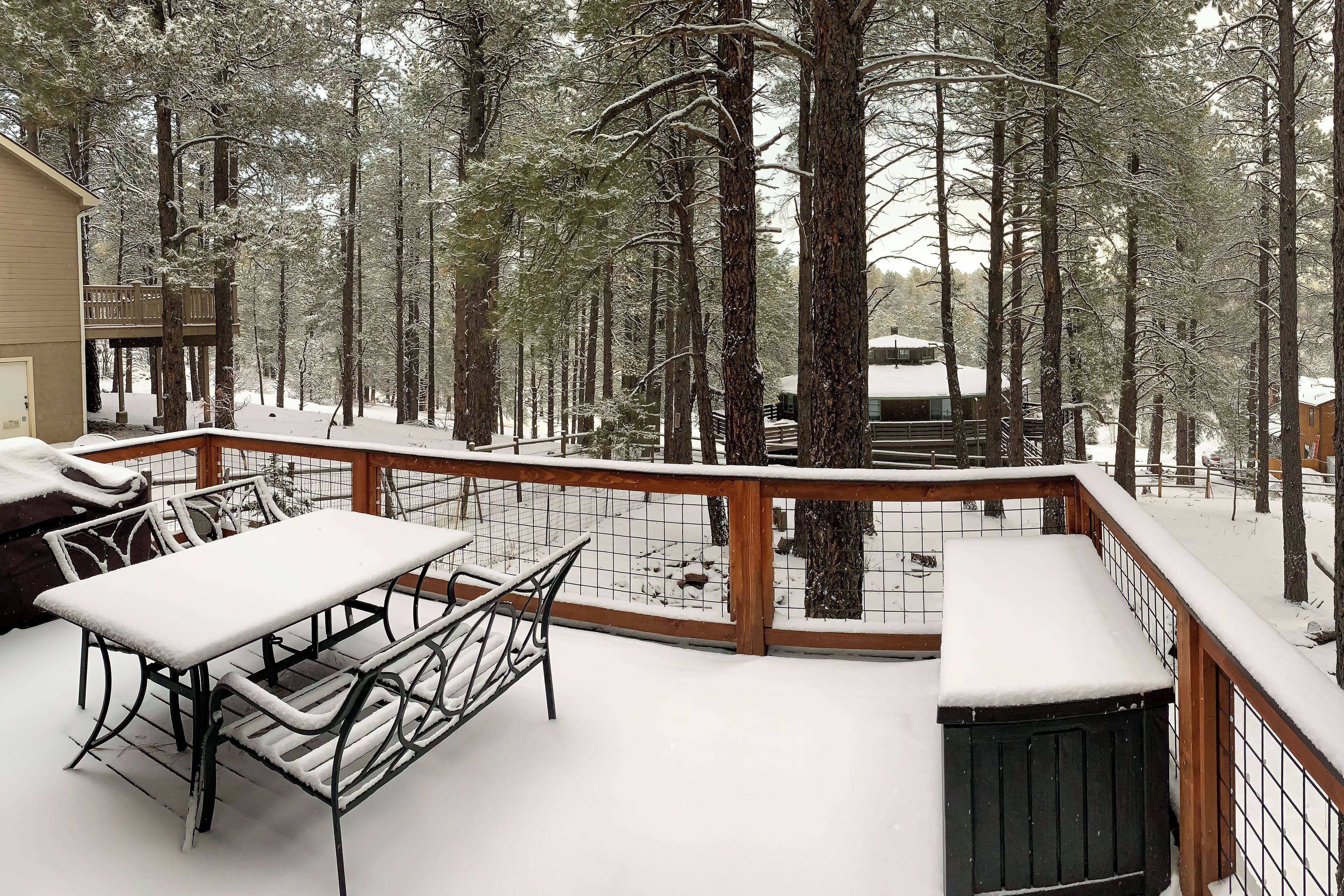 A snow-covered deck with a table and benches, surrounded by tall trees.