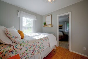Un dormitorio con cama, ventana con persianas, mesita de noche con planta y puerta que da a otra habitación.