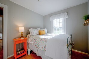 Quarto com cama, mesa de cabeceira, abajur, janela com persianas e prateleira pequena com uma planta.