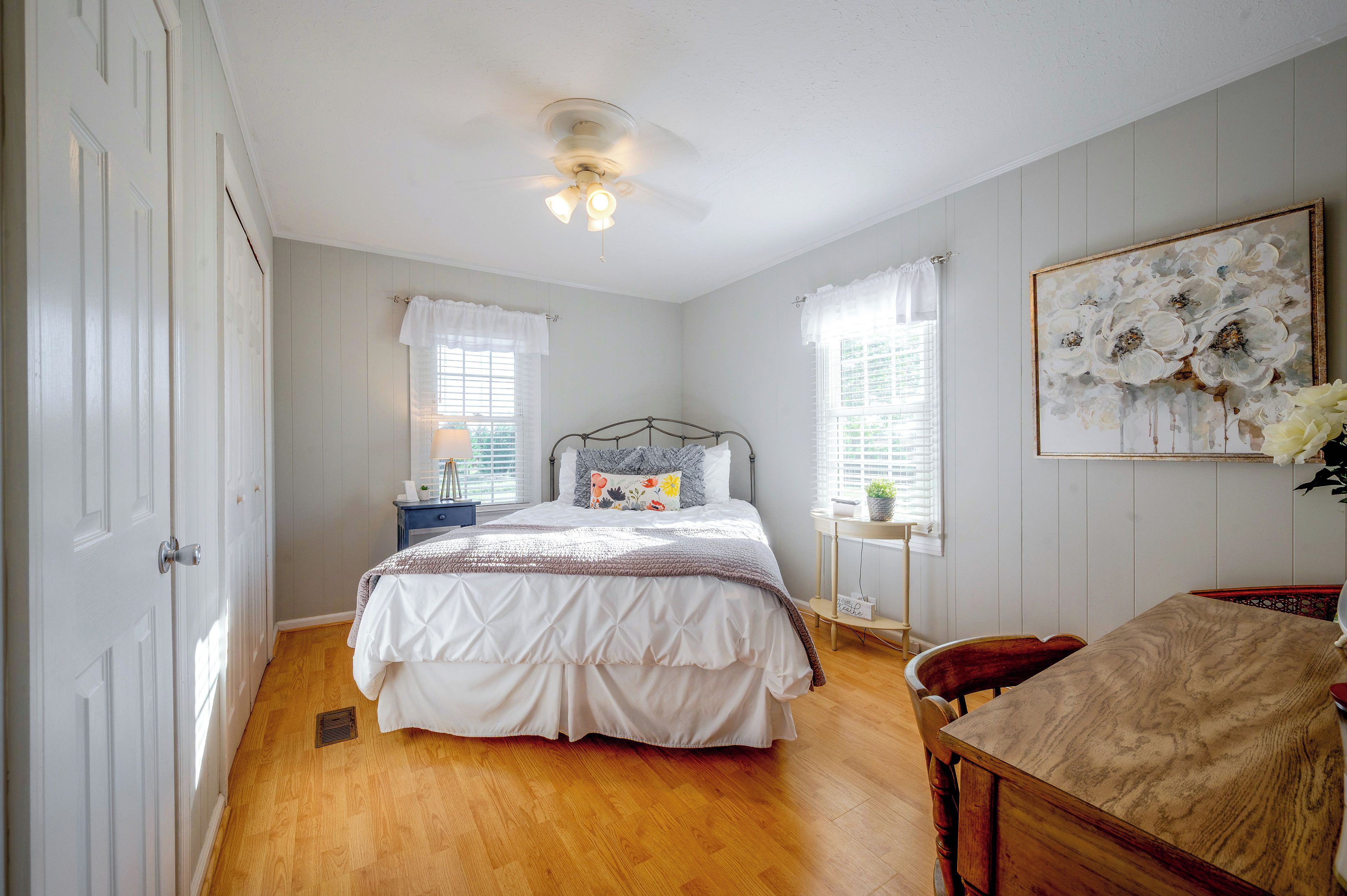 Une chambre à coucher comprenant un lit, des tables de chevet, une chaise, un ventilateur de plafond et une fenêtre avec des rideaux.
