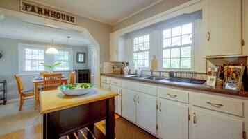 A kitchen with white cabinets, a farmhouse sign, a dining table with chairs, and a window with a view of trees.