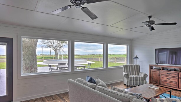 A spacious living room with a large window, a ceiling fan, a television, and a wooden coffee table. - Fire Pit, Pickleball, Pool! Schulenburg Farmhouse (Schulenburg)